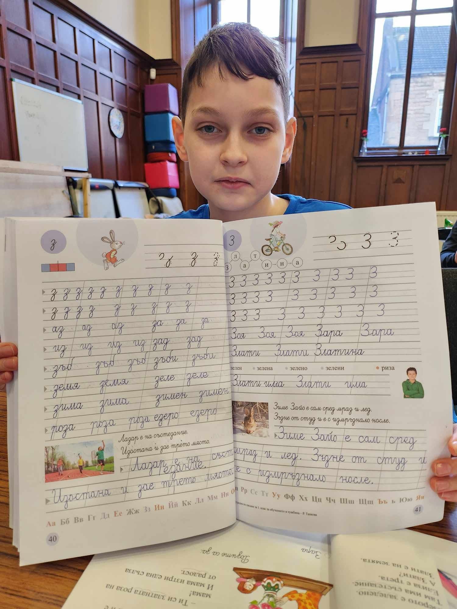 A pupil holding up his Bulgarian handwriting workbook, open at exercises on the letter “з”